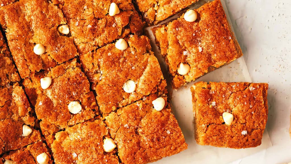 A batch of blondies with white chocolate chips is cut into squares and arranged on parchment paper like a sheet cake recipe come to life. One square is slightly separated from the others. The blondies have a golden-brown crust and are lightly sprinkled with sea salt.