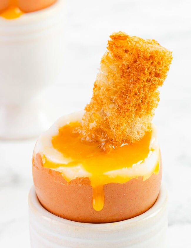 Breakfast delight: Soft-boiled egg in an egg cup with runny yolk and a piece of toasted bread for dipping. Another egg cup and an empty half eggshell are in the background, all on a marble surface, perfect for easy egg recipes that make your morning special.