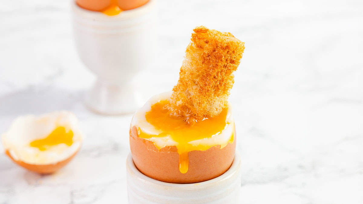 Breakfast delight: Soft-boiled egg in an egg cup with runny yolk and a piece of toasted bread for dipping. Another egg cup and an empty half eggshell are in the background, all on a marble surface, perfect for easy egg recipes that make your morning special.