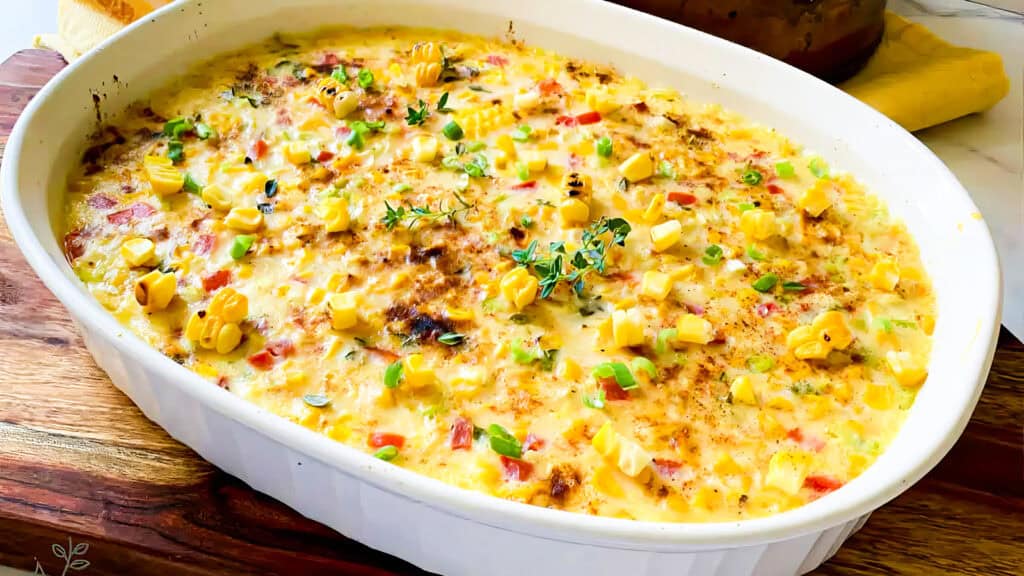 A white baking dish filled with a baked casserole topped with corn, bell peppers, and green onions, garnished with fresh herbs. The dish embodies the essence of Southern recipes, resting on a wooden surface with a yellow napkin in the background.