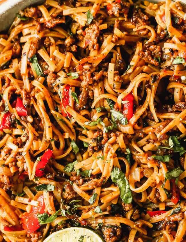 A bowl of stir-fried noodles garnished with slices of lime. The dish contains noodles mixed with various vegetables, herbs, ground beef, and pieces of red bell pepper. Chopsticks are placed beside the bowl on a marble surface.