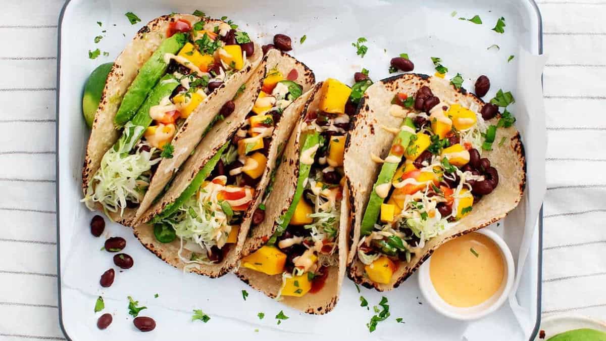 A tray holding four tacos filled with vibrant ingredients including lettuce, diced mango, black beans, fresh avocados slices, chopped cilantro, and a drizzle of creamy sauce. A small bowl filled with orange sauce sits on the side. The tacos are presented on a white platter.