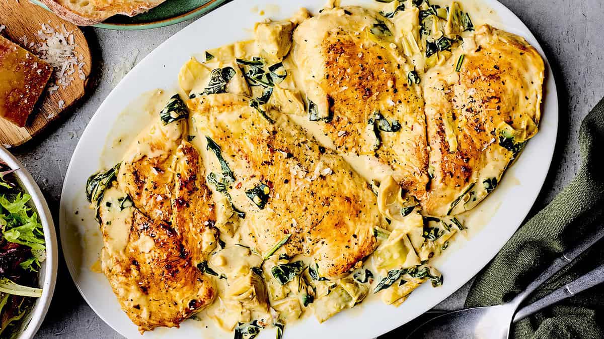 A plate of creamy garlic chicken is presented, featuring golden-brown chicken breasts topped with a rich, creamy sauce that contains spinach and artichokes. This easy peasy dish is garnished with cracked black pepper and sits on a gray countertop with a green napkin nearby—perfect for back-to-school recipes.