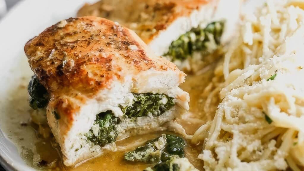 A close-up of a plate featuring a stuffed chicken breast filled with spinach and cheese, showcasing one of the tastiest spinach recipes. It is sliced open to reveal the filling. Next to the chicken is a serving of creamy pasta garnished with grated cheese.