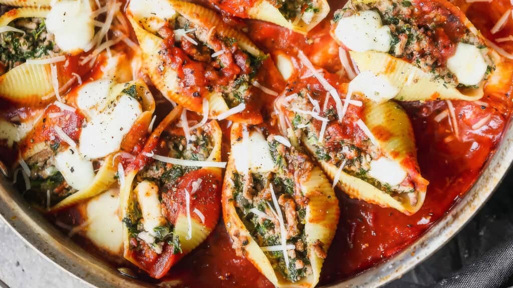 A close-up of a dish featuring large pasta shells stuffed with a mixture of ground beef and spinach, generously covered in tomato sauce and sprinkled with shredded cheese. The dish appears baked and garnished with fresh herbs and grated parmesan, making it a standout among ground beef recipes.