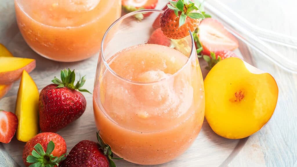 A glass of refreshing peach and strawberry smoothie garnished with a fresh strawberry on the rim, surrounded by whole strawberries, sliced mango, and peach halves on a light wooden surface. Another smoothie glass is partially visible in the background, offering inspiration for cocktail recipes.