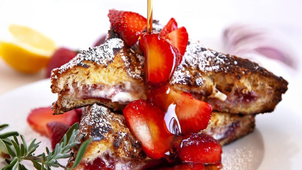 A close-up of a plate with French toast stuffed with a creamy filling and topped with fresh strawberry slices. Powdered sugar is lightly dusted on top, and syrup is being drizzled from above. Blurred in the background, a lemon wedge and rosemary sprig add elegance reminiscent of gourmet pancake recipes.