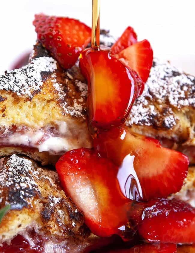 A close-up of a plate with French toast stuffed with a creamy filling and topped with fresh strawberry slices. Powdered sugar is lightly dusted on top, and syrup is being drizzled from above. Blurred in the background, a lemon wedge and rosemary sprig add elegance reminiscent of gourmet pancake recipes.