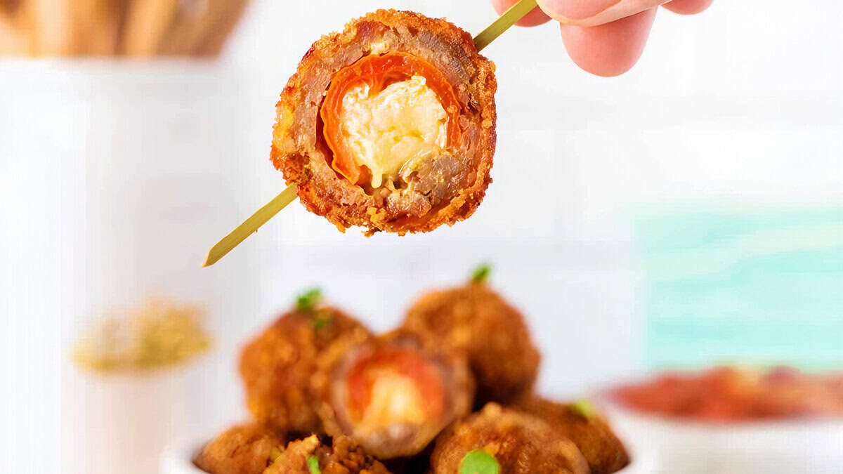 A hand holds a skewer with a bite-sized breaded and fried meatball, revealing a filling of melted cheese and a cherry tomato. More of the same delectable appetizers, perfect for any cooking book focused on meatball dishes, are visible in a bowl in the soft-focus background.