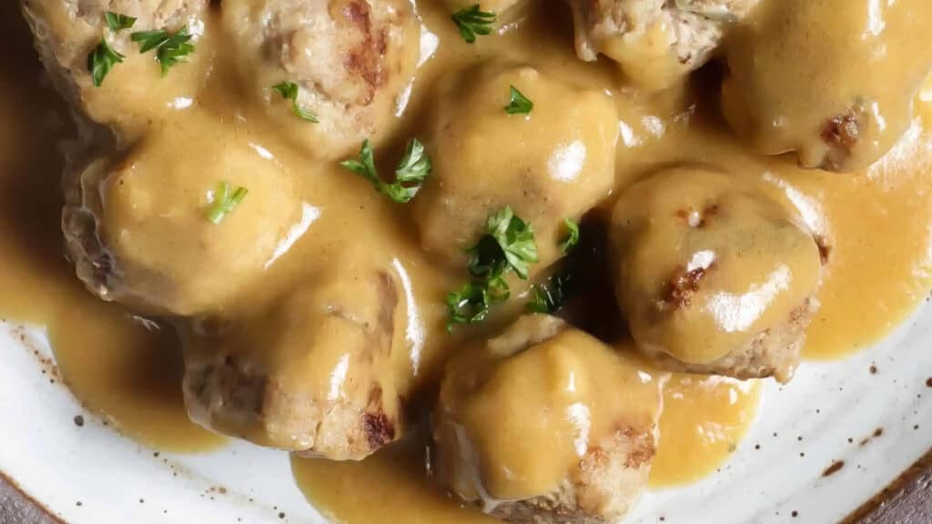 A plate of Swedish meatballs covered in creamy brown gravy and garnished with chopped parsley. The ground beef recipe ensures the meatballs are tender and generously coated with thick, rich gravy, giving a hearty and comforting appearance.