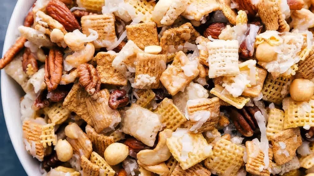 A close-up view of a snack mix consisting of cereal squares, pretzel pieces, roasted nuts, and coconut flakes. This delightful blend from your favorite snack recipes appears to be lightly coated with a sweet glaze, giving it a glossy appearance.