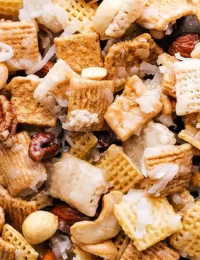 A close-up view of a snack mix consisting of cereal squares, pretzel pieces, roasted nuts, and coconut flakes. This delightful blend from your favorite snack recipes appears to be lightly coated with a sweet glaze, giving it a glossy appearance.