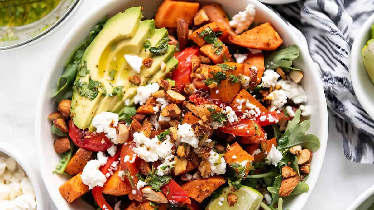 A vibrant salad in a white bowl features nutrient-rich sliced avocado, roasted sweet potatoes, red bell peppers, arugula, crumbled feta cheese, chopped almonds, and a sprinkle of fresh herbs, all drizzled with a light dressing. A striped napkin is partially visible in the background.