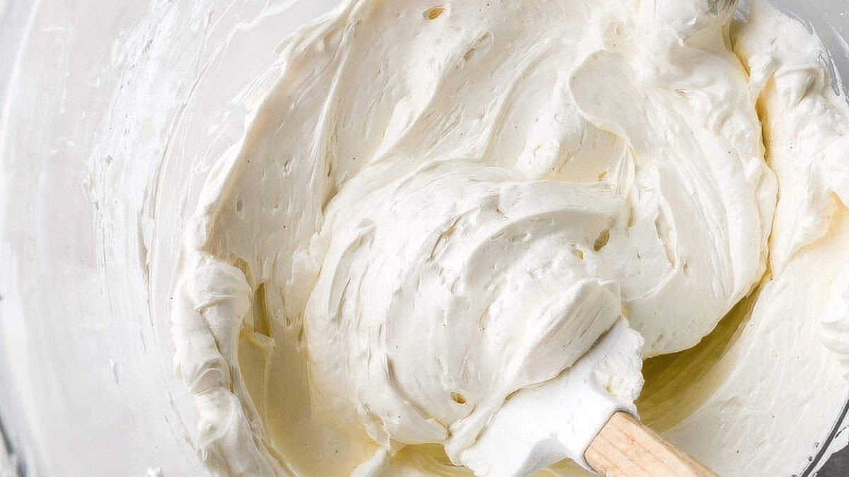 A close-up of a mixing bowl filled with creamy, light-colored batter, perfect for Cakes or Cupcakes. A white spatula is partially submerged in the batter, with some of the mixture clinging to it. The texture appears smooth and whipped, ideal for various frosting recipes.