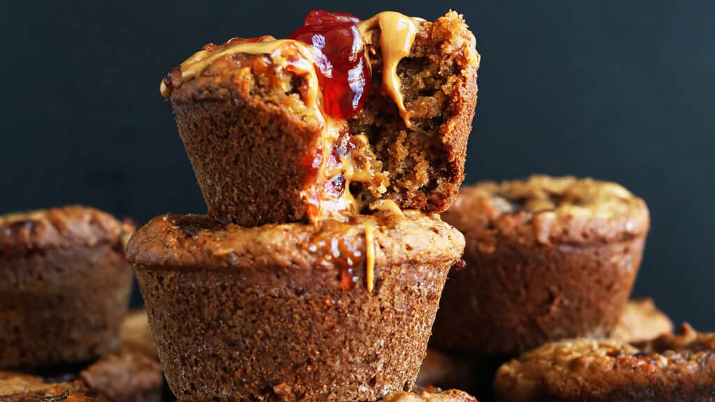 A stack of Peanut Butter and Jelly Muffins, with one muffin on top cut open, revealing a filling of peanut butter and jelly oozing out. The muffins have a slightly rough texture and appear fresh and moist, set against a dark background—ideal for those exploring new muffin recipes.