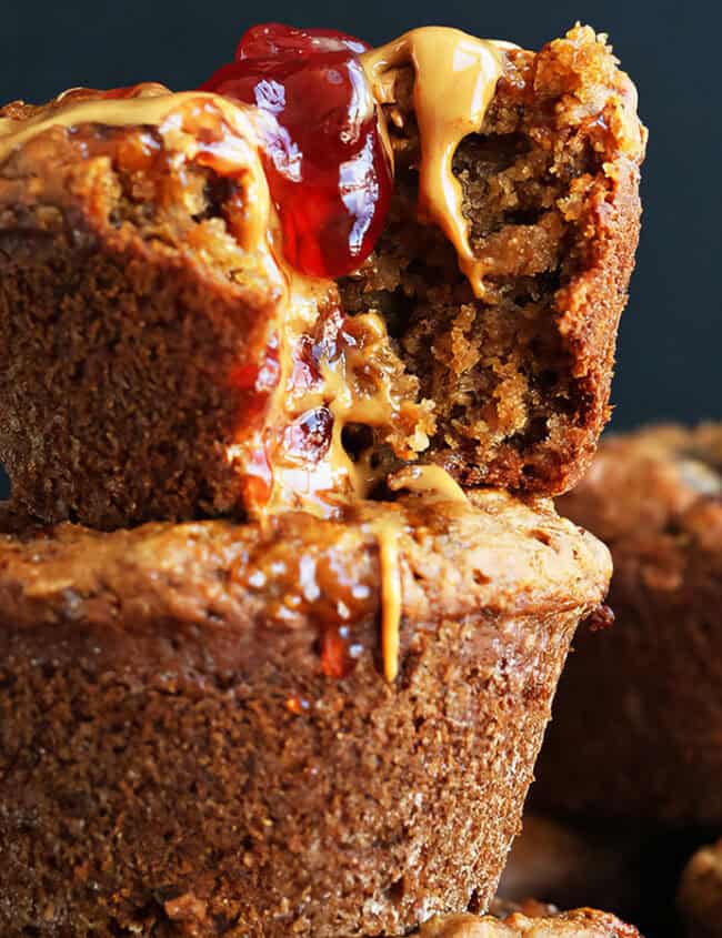 A stack of Peanut Butter and Jelly Muffins, with one muffin on top cut open, revealing a filling of peanut butter and jelly oozing out. The muffins have a slightly rough texture and appear fresh and moist, set against a dark background—ideal for those exploring new muffin recipes.