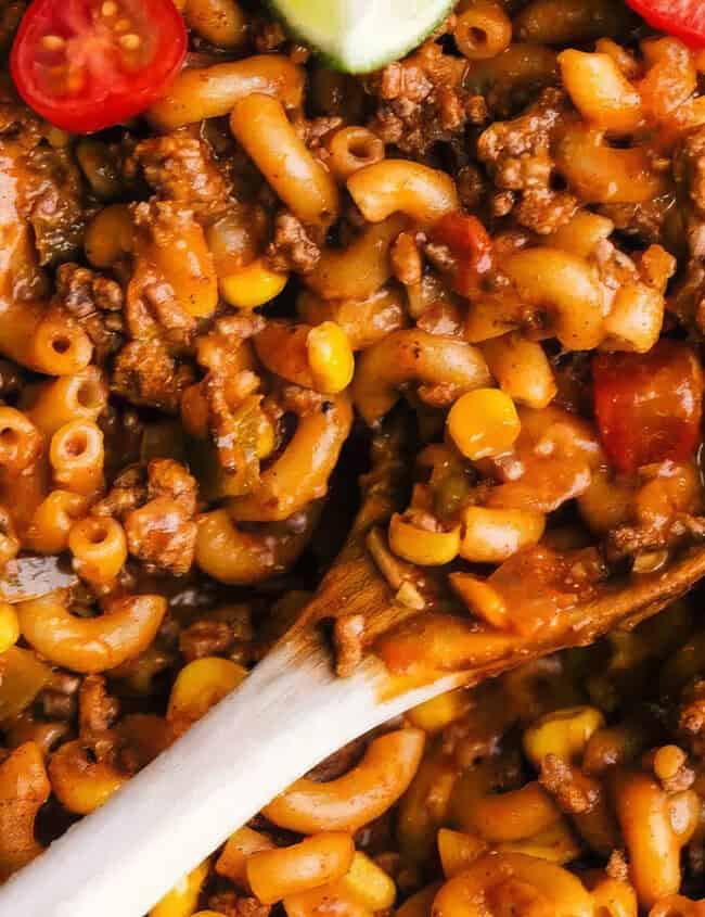 A close-up of a hearty pasta dish filled with macaroni noodles, ground meat, corn, and slices of cherry tomatoes. A wooden spoon is seen stirring the mixture, which is covered in a rich, tomato-based sauce. Perfect for back-to-school recipes, this easy dish will delight the whole family.