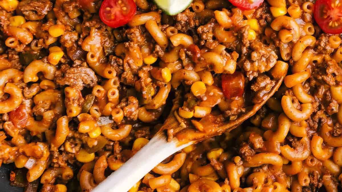 A close-up of a hearty pasta dish filled with macaroni noodles, ground meat, corn, and slices of cherry tomatoes. A wooden spoon is seen stirring the mixture, which is covered in a rich, tomato-based sauce. Perfect for back-to-school recipes, this easy dish will delight the whole family.