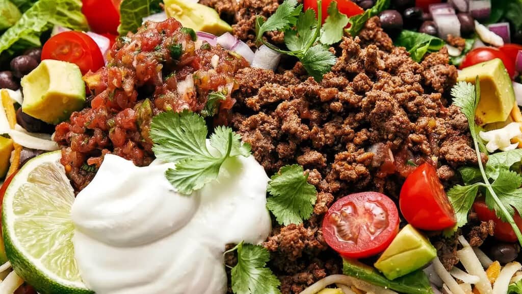 A close-up of a colorful taco salad topped with ground beef, sour cream, salsa, cilantro, chopped tomatoes, avocado, and a lime wedge. This delicious dish is a standout among ground beef recipes, with its mix of ingredients creating a vibrant and appetizing appearance.