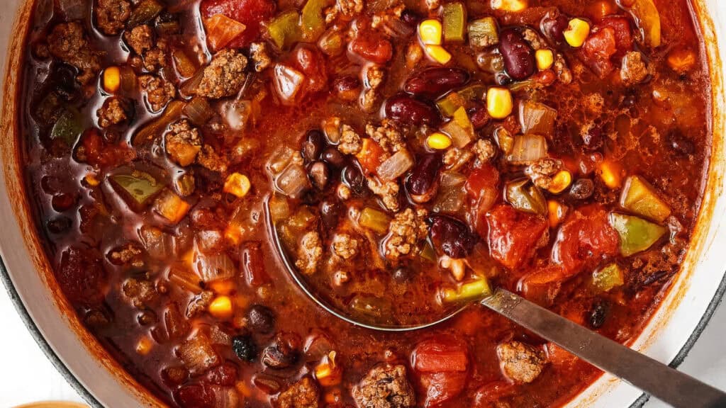 A close-up of a pot of chili, showcasing one of the best ground beef recipes. The rich, thick stew is filled with kidney beans, black beans, corn, diced green bell peppers, tomatoes, and onions. A ladle is partially submerged in the chili, highlighting the chunky ingredients and vibrant colors.