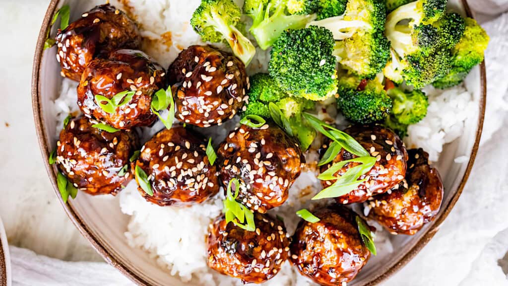 A bowl filled with white rice, glazed meatballs sprinkled with sesame seeds and chopped green onions, and a portion of steamed broccoli. The presentation showcases a colorful and appetizing dish that could easily be featured among 20 top recipes for a quick and delicious meal.