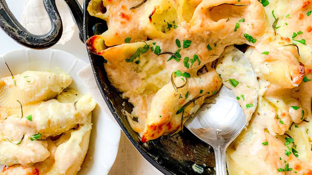 A close-up of a baked pasta dish with large shell pasta stuffed with creamy cheese, topped with melted cheese and sprinkled with chopped herbs. A silver spoon rests in the dish, ready for serving. A portion of the pasta is served on a white plate nearby, delighting all pasta lovers.