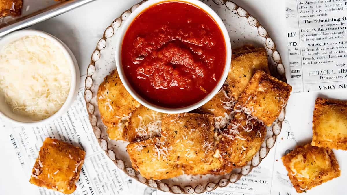 A plate of golden-brown stuffed pasta topped with grated cheese surrounds a bowl of marinara sauce. The plate rests on a scattered, rustic collection of newspaper pages. A small bowl of grated cheese is seen beside the plate—a perfect treat for Pasta Lovers.