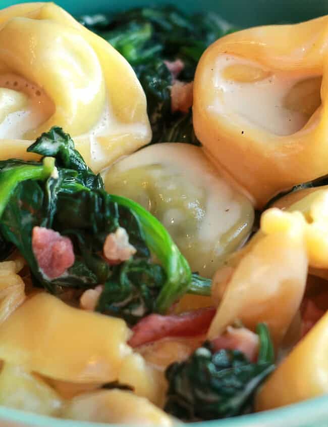 A green bowl filled with tortellini pasta in a creamy Alfredo sauce, mixed with wilted spinach and small pieces of bacon. The dish appears rich and savory, promising to be a good indulgence for any meal.