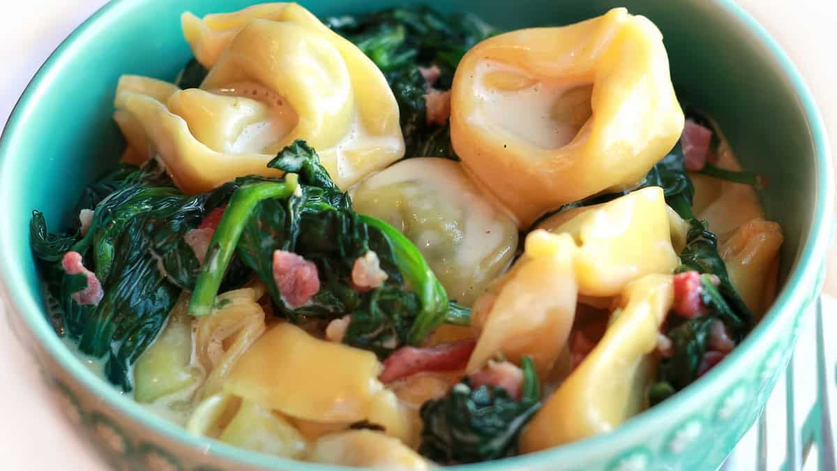 A green bowl filled with tortellini pasta in a creamy Alfredo sauce, mixed with wilted spinach and small pieces of bacon. The dish appears rich and savory, promising to be a good indulgence for any meal.