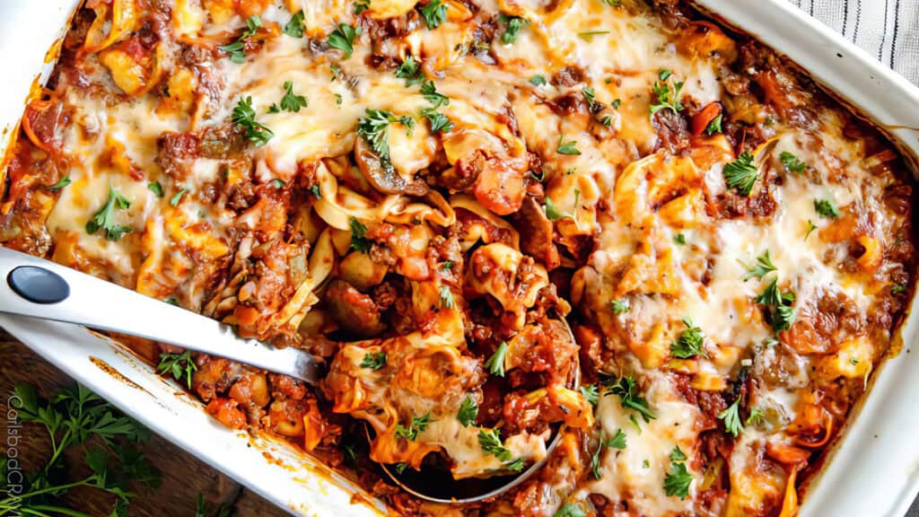 A close-up of a baked cheesy lasagna in a rectangular white dish. The lasagna, one of the best ground beef recipes, is topped with melted cheese and garnished with fresh parsley. A serving spoon is partially scooping out a portion, revealing layers of pasta, sauce, and ground meat inside.