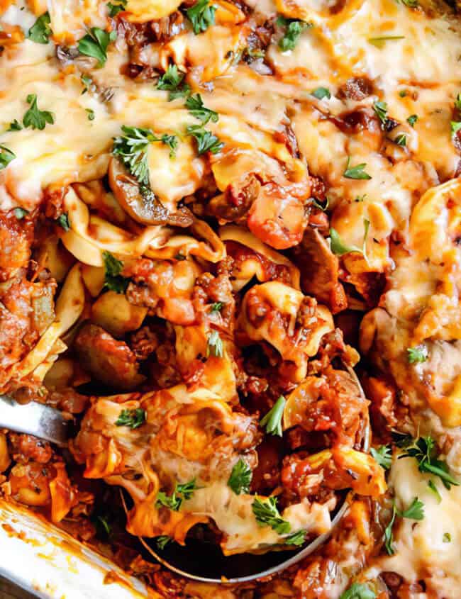 A close-up of a baked cheesy lasagna in a rectangular white dish. The lasagna, one of the best ground beef recipes, is topped with melted cheese and garnished with fresh parsley. A serving spoon is partially scooping out a portion, revealing layers of pasta, sauce, and ground meat inside.