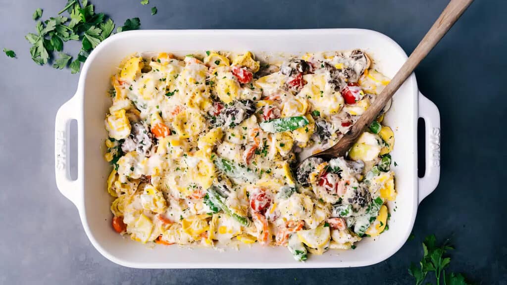 A rectangular white baking dish filled with a colorful and cheesy pasta bake, consisting of tortellini, cherry tomatoes, spinach, mushrooms, and creamy sauce. A wooden spoon rests in the dish, with garnished herbs visible on top. Fresh parsley is scattered nearby—a perfect addition to your casserole recipes.