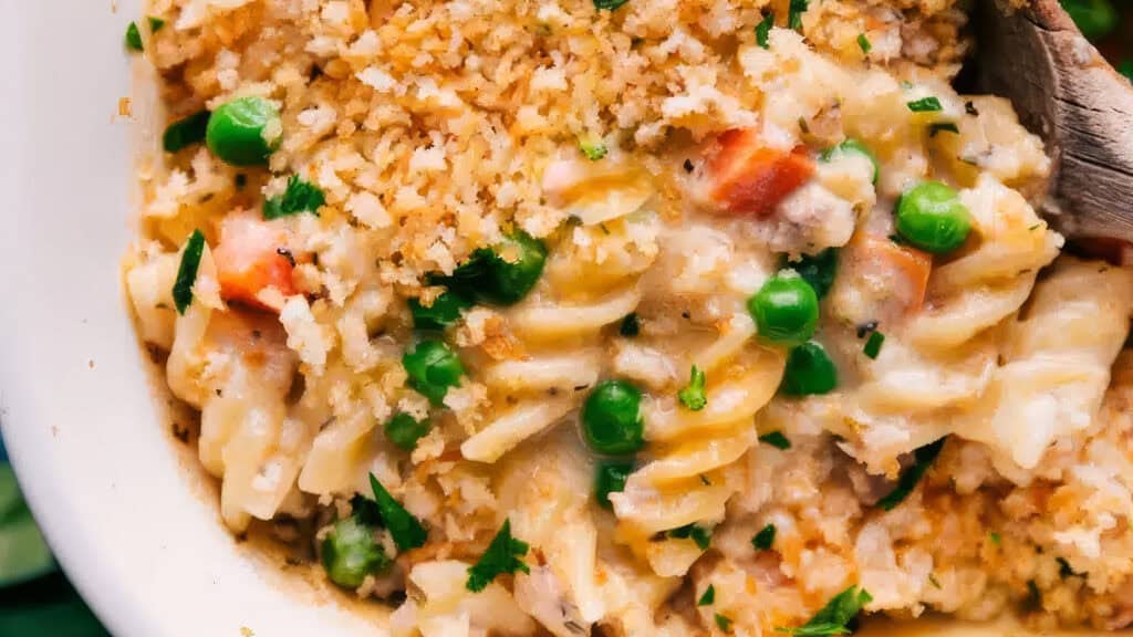 Close-up of a creamy pasta bake with rotini noodles, green peas, diced carrots, and a golden breadcrumb topping. The dish is garnished with chopped parsley, adding a touch of color to the rich casserole. Perfect for those who love hearty casserole recipes. A wooden spoon is partially visible on the right side.