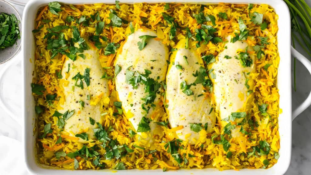 A rectangular white baking dish contains a vibrant baked meal with four pieces of white fish fillets on a bed of yellow rice. The rice, part of classic rice dish recipes, is garnished with chopped green herbs, giving it a colorful and fresh appearance.