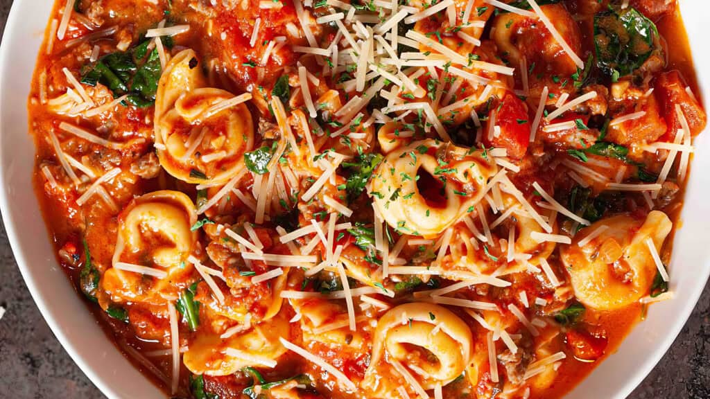 A close-up of a bowl filled with tortellini in a rich tomato sauce, garnished with shredded parmesan cheese and chopped parsley. The dish is vibrant, showcasing one of the finest spinach recipes, with visible chunks of tomatoes and pieces of fresh spinach mixed throughout.