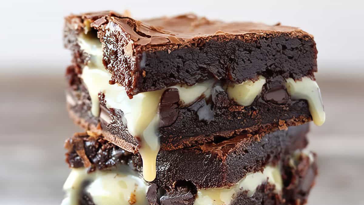 A close-up shot of two stacked, rich chocolate brownies with a glossy top crust and gooey, melty white chocolate filling oozing out. These brownies go beyond basic, tantalizing chocoholics with their crispy exterior and luscious, creamy interior. The background is blurred.