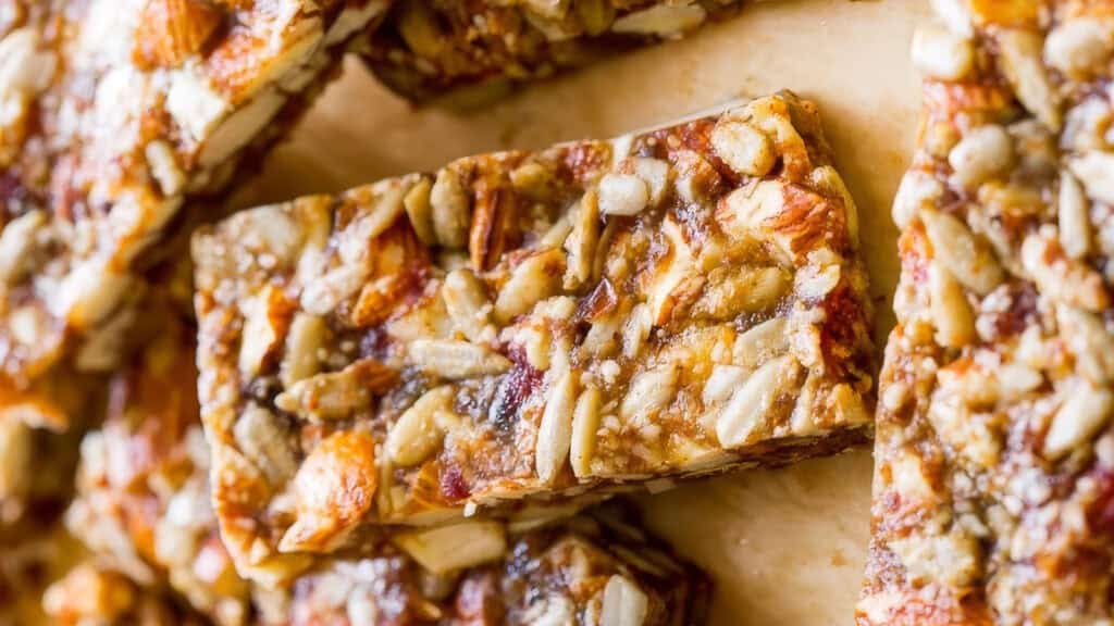 Close-up of several nut and seed bars on a light-colored surface. These delightful treats, straight from snack recipes, are made with various nuts and seeds bound together, showcasing a glossy and crunchy texture.