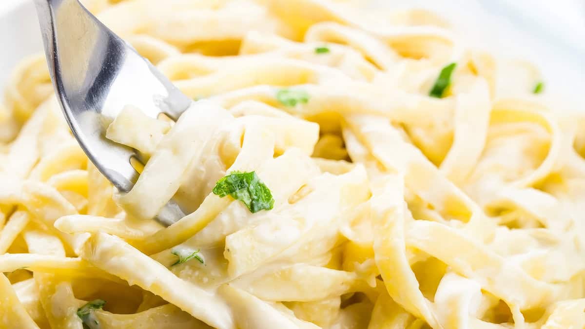 A close-up of a fork twirling creamy Alfredo fettuccine pasta garnished with chopped parsley. The sauce is thick and velvety, coating the pasta evenly—truly one of the good Alfredo recipes. The background is a light blur, emphasizing the dish's rich texture and inviting appearance.