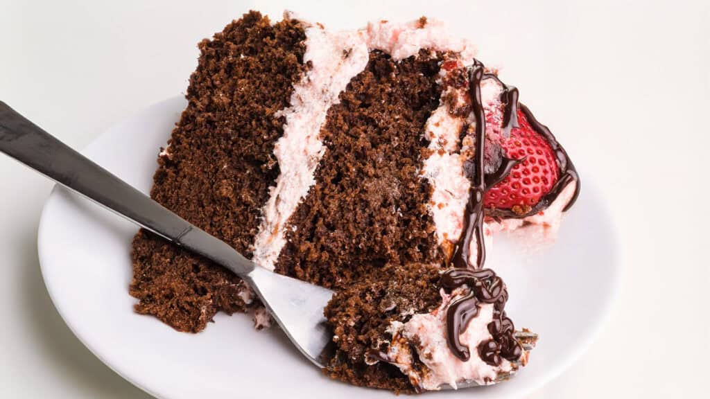 A slice of rich chocolate cake layered with light pink frosting is served on a white plate. As one of the finest strawberry desserts, a fresh strawberry sits beside the cake, drizzled with chocolate syrup. A fork is partially inserted into the cake, ready to take a bite.