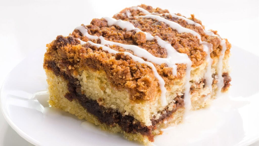 A close-up of an irresistible coffee cake slice on a white plate. The cake features a moist, golden interior layered with a cinnamon-brown sugar filling and topped with a crispy crumb topping. Wake up and smell the coffee cake as it's drizzled with a light glaze.