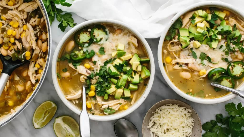 Three bowls of hearty chicken and white bean soup, a delightful addition to your one pot recipes collection, are garnished with chopped avocado, cheese, cilantro, and jalapenos. Beside them sits a pot of the soup, fresh lime wedges, a white towel, and a bowl of shredded cheese on the white surface.