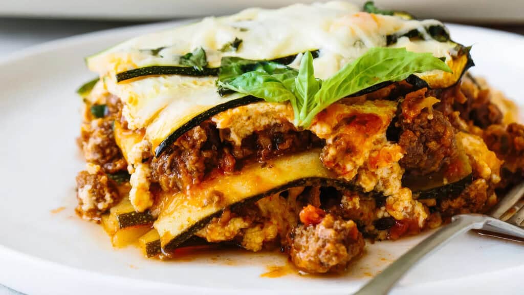 A slice of zucchini lasagna is presented on a white plate with a fork beside it. This delicious entry in ground beef recipes features layers of zucchini, ground meat, tomato sauce, and melted cheese, all garnished with a fresh basil leaf on top.