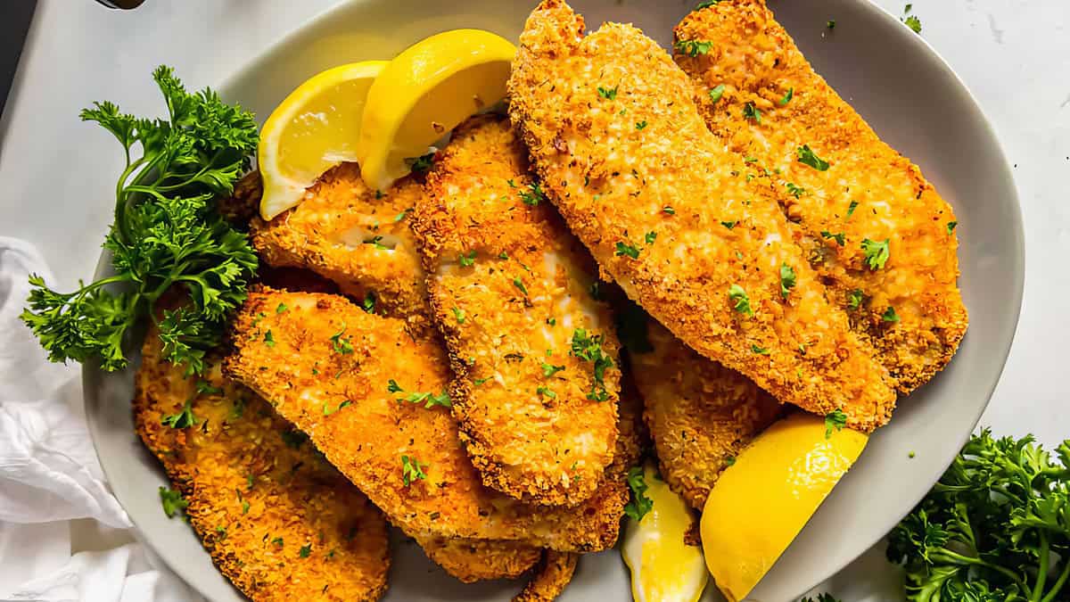 A plate filled with crispy, golden-breaded fish fillets garnished with chopped parsley. The fish is accompanied by fresh parsley sprigs and lemon wedges for added flavor. The background includes a white cloth and a few parsley sprigs, making it perfect for breaded recipes enthusiasts.