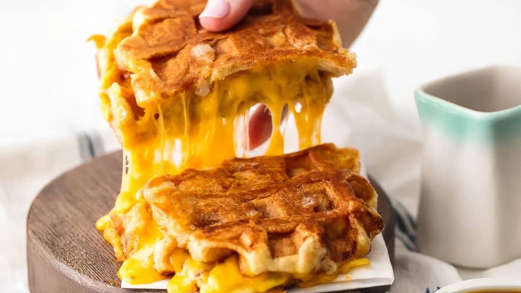 A close-up of a hand pulling apart two cheesy waffle sandwiches, showing gooey melted cheese stretching between the golden, crispy waffles. A small white jug with a mint green rim, often seen in pancake recipes, is partially visible on the side.