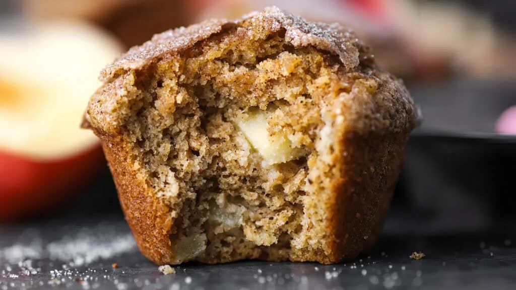 Close-up of a bitten apple cinnamon muffin. The interior displays a moist, crumbly texture with visible chunks of apple. The muffin top is slightly sugary, creating a contrast with the soft inside. The background is blurred with hints of other muffins, perfect inspiration for new muffin recipes.