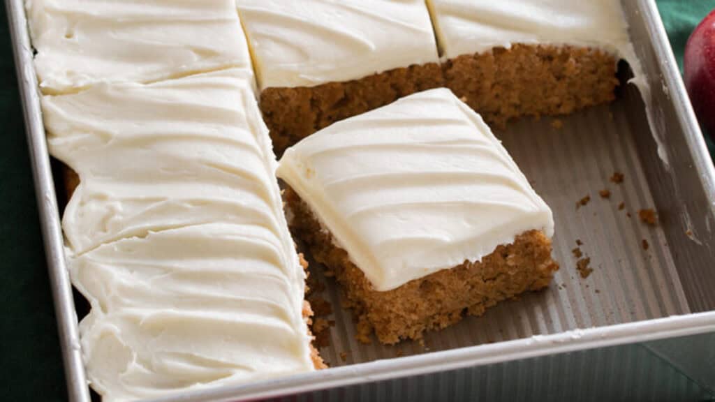 A tray of frosted rectangular cake slices, with one piece slightly removed from the pan, showcases a delectable example from sheet cake recipes. The moist cake boasts a thick layer of smooth white icing on top, and the metallic tray features a ridged texture at the base. Some apple pieces are visible at the edge.