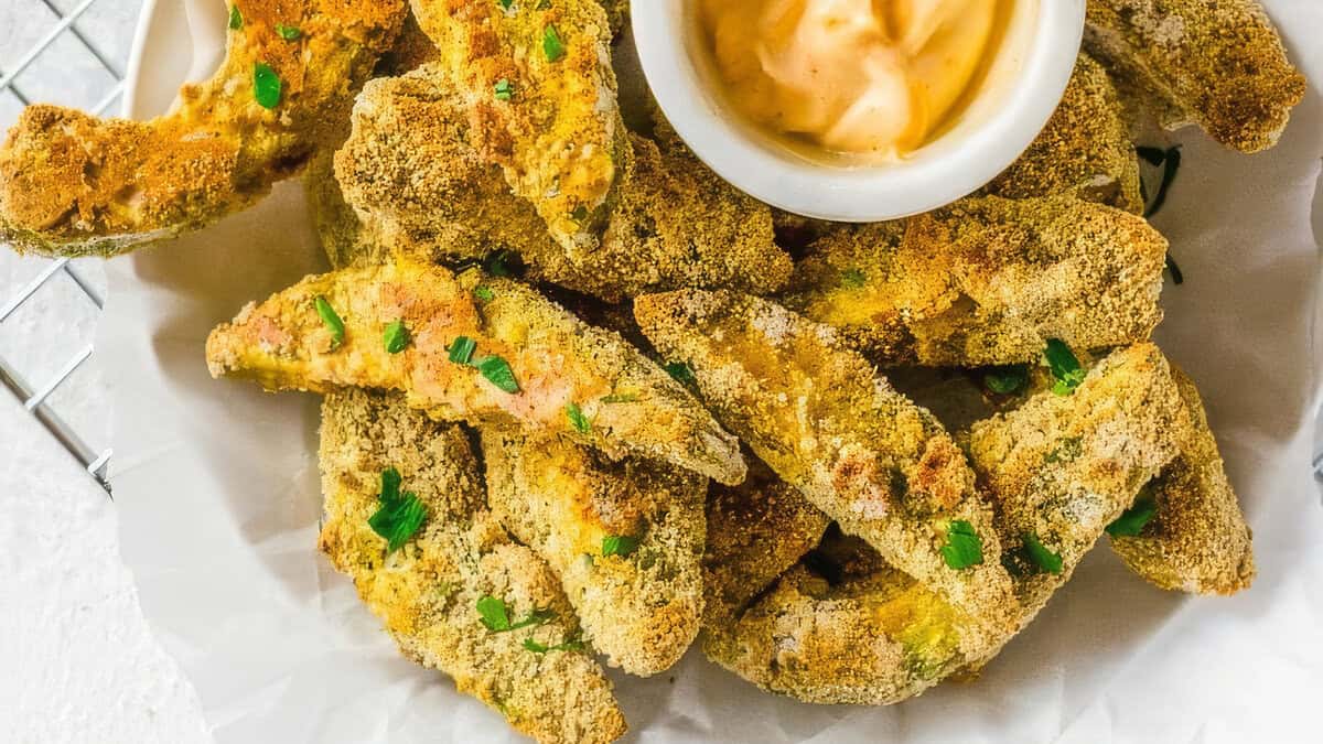 A plate of crispy, golden-brown breaded avocado fries, garnished with chopped herbs, is served with a small bowl of creamy dipping sauce. The healthy avocado fries are arranged on white parchment paper, creating an appetizing presentation.