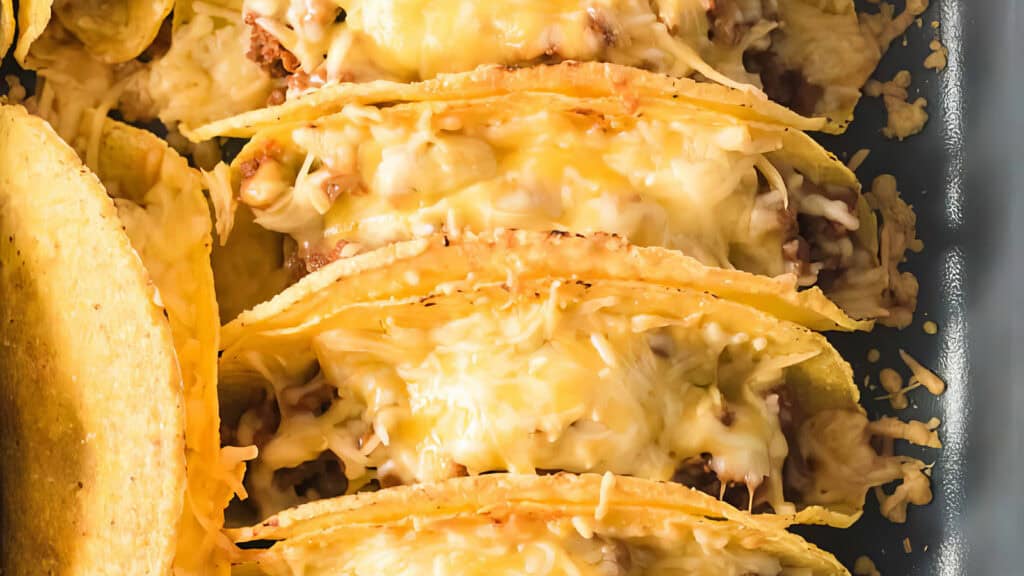 A close-up view of a baking tray filled with hard-shell tacos, generously topped with melted cheese. These delicious ground beef recipes are packed closely together and arranged in rows. The melted cheese is golden and bubbly, covering the filling and shell edges.