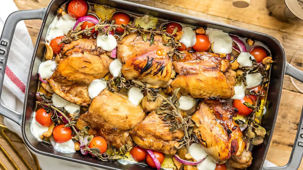 A roasted chicken dish in a rectangular baking pan. The pan contains browned chicken pieces, cherry tomatoes, red onion slices, garlic cloves, and herbs, all topped with dollops of melted white cheese. Drawing inspiration from Mediterranean recipes, the pan rests on a wooden surface with a red-striped towel beside it.