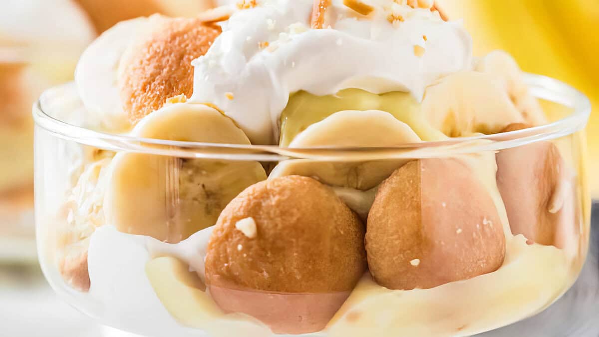 A close-up of a dessert bowl containing banana slices, vanilla pudding, round wafer cookies, and whipped cream. This Southern recipe is topped with a garnish of crushed nuts or cookie crumbs. The background is softly blurred, highlighting the dessert's textures and layers.